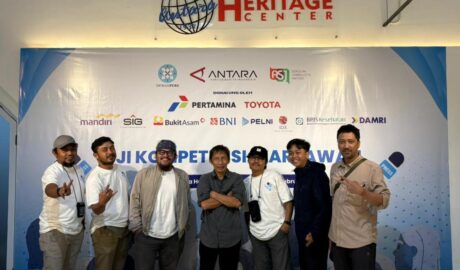 Tujuh pria berdiri berjejer di depan latar belakang backdrop bertuliskan "Uji Kompetensi Wartawan" dengan logo berbagai sponsor. Mereka berpose kasual dan menunjukkan ekspresi senang setelah menyelesaikan ujian.