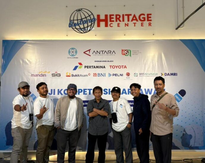 Tujuh pria berdiri berjejer di depan latar belakang backdrop bertuliskan "Uji Kompetensi Wartawan" dengan logo berbagai sponsor. Mereka berpose kasual dan menunjukkan ekspresi senang setelah menyelesaikan ujian.