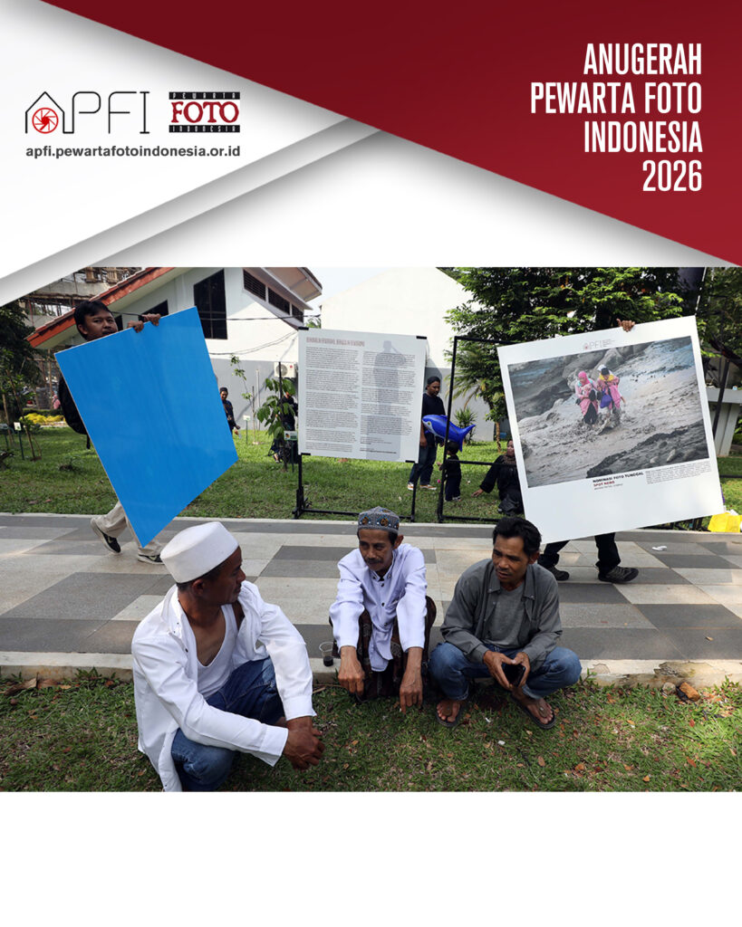 Tiga warga lokal duduk berbincang santai di tepi rumput, berlatar belakang kesibukan anggota panitia yang berlalu-lalang menggotong panel foto berukuran besar.