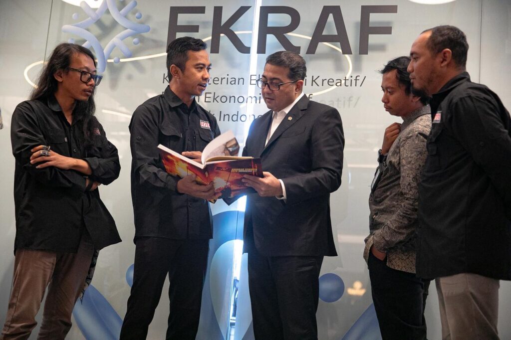 Menekraf Teuku Riefky Harsya bersama pengurus PFI berdiri melingkar mencermati isi halaman cetak Buku Foto APFI 2026 di depan dinding kaca bertuliskan EKRAF.