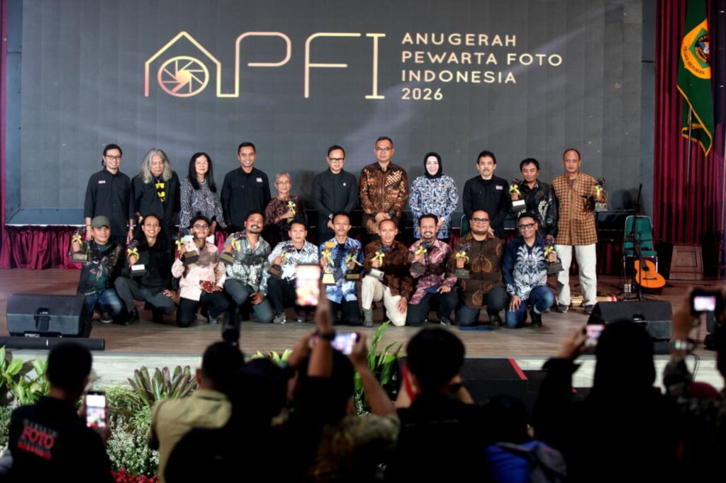 Momen penganugerahan gelar tertinggi Photo Of The Year APFI 2026 kepada Fauzan di atas panggung Auditorium Sekretariat Daerah Kabupaten Bogor, 8 Mei 2026. Fauzan berdiri di podium berbicara ke mikrofon sambil memegang bingkai foto besar karyanya yang monumental. Di sampingnya berdiri pria berkemeja batik memegang trofi. Backdrop LED besar di belakang mereka bertuliskan 'APFI 2026', 'Photo Of The Year', 'Fauzan', 'Demonstrasi Atas Tewasnya Ojol Yang Terlindas Rantis Brimob', dan logo PFI Pusat serta Pemkab Bogor.
