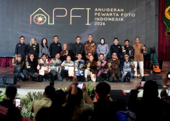 Momen penganugerahan gelar tertinggi Photo Of The Year APFI 2026 kepada Fauzan di atas panggung Auditorium Sekretariat Daerah Kabupaten Bogor, 8 Mei 2026. Fauzan berdiri di podium berbicara ke mikrofon sambil memegang bingkai foto besar karyanya yang monumental. Di sampingnya berdiri pria berkemeja batik memegang trofi. Backdrop LED besar di belakang mereka bertuliskan 'APFI 2026', 'Photo Of The Year', 'Fauzan', 'Demonstrasi Atas Tewasnya Ojol Yang Terlindas Rantis Brimob', dan logo PFI Pusat serta Pemkab Bogor.