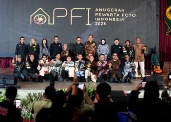 Momen penganugerahan gelar tertinggi Photo Of The Year APFI 2026 kepada Fauzan di atas panggung Auditorium Sekretariat Daerah Kabupaten Bogor, 8 Mei 2026. Fauzan berdiri di podium berbicara ke mikrofon sambil memegang bingkai foto besar karyanya yang monumental. Di sampingnya berdiri pria berkemeja batik memegang trofi. Backdrop LED besar di belakang mereka bertuliskan 'APFI 2026', 'Photo Of The Year', 'Fauzan', 'Demonstrasi Atas Tewasnya Ojol Yang Terlindas Rantis Brimob', dan logo PFI Pusat serta Pemkab Bogor.