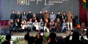 Momen penganugerahan gelar tertinggi Photo Of The Year APFI 2026 kepada Fauzan di atas panggung Auditorium Sekretariat Daerah Kabupaten Bogor, 8 Mei 2026. Fauzan berdiri di podium berbicara ke mikrofon sambil memegang bingkai foto besar karyanya yang monumental. Di sampingnya berdiri pria berkemeja batik memegang trofi. Backdrop LED besar di belakang mereka bertuliskan 'APFI 2026', 'Photo Of The Year', 'Fauzan', 'Demonstrasi Atas Tewasnya Ojol Yang Terlindas Rantis Brimob', dan logo PFI Pusat serta Pemkab Bogor.