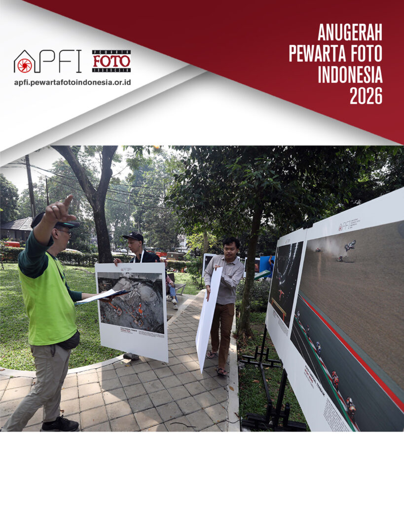 Panitia berrompi hijau memberikan aba-aba penempatan lokasi pameran kepada dua relawan pemuda yang menggotong panel-panel foto besar berisi dokumentasi gempa dan trek balap.