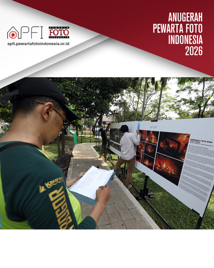 Bidikan dari sudut belakang bahu panitia berkaos hijau bertuliskan "BOGOR" yang sedang memegang papan klip daftar periksa, memantau pemasangan panel foto esai di taman.