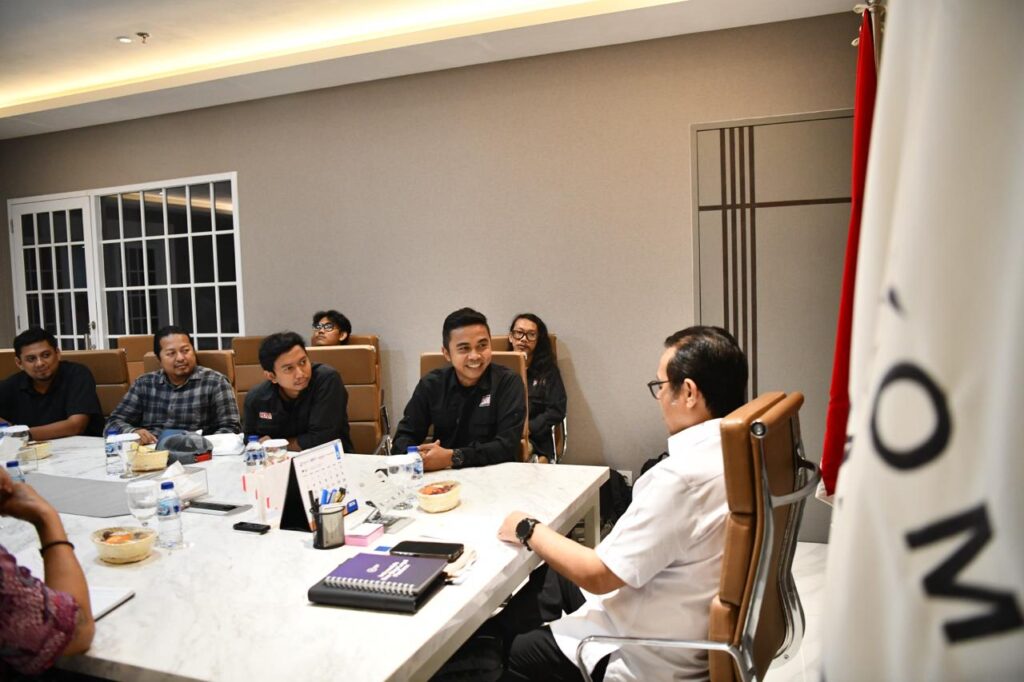 Sudut pandang kamera dari samping belakang Wamen Komdigi memperlihatkan senyum cerah dan cair para pengurus PFI saat berdiskusi di ruang rapat.