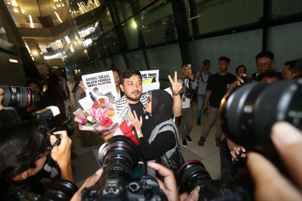 Thoudy Badai memegang buket bunga dikerumuni fotografer saat dipeluk erat oleh istrinya yang menangis haru di terminal kedatangan bandara.